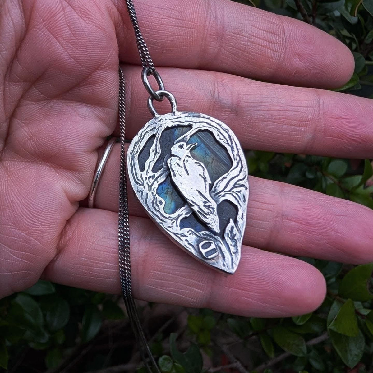 Ciondolo Il Bosco del Corvo in argento 925: corvo tra i rami su labradorite, a goccia, sul palmo della mano