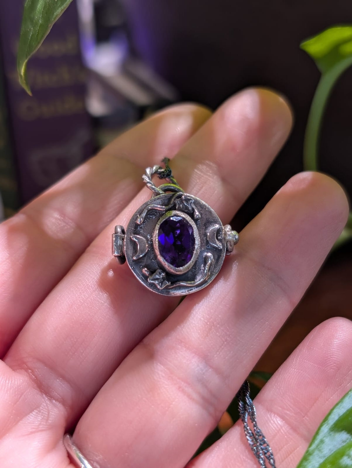 The Witch's Secret locket closed, in burnished 925 silver with a faceted purple amethyst, ivy sprigs and crescent moons, resting on a hand